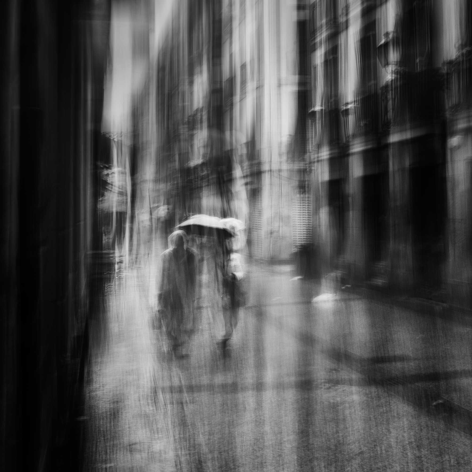 Black and white image of blurred figures under an umbrella on a rainy street in San Sebastian, Spain, creating an ethereal atmosphere.