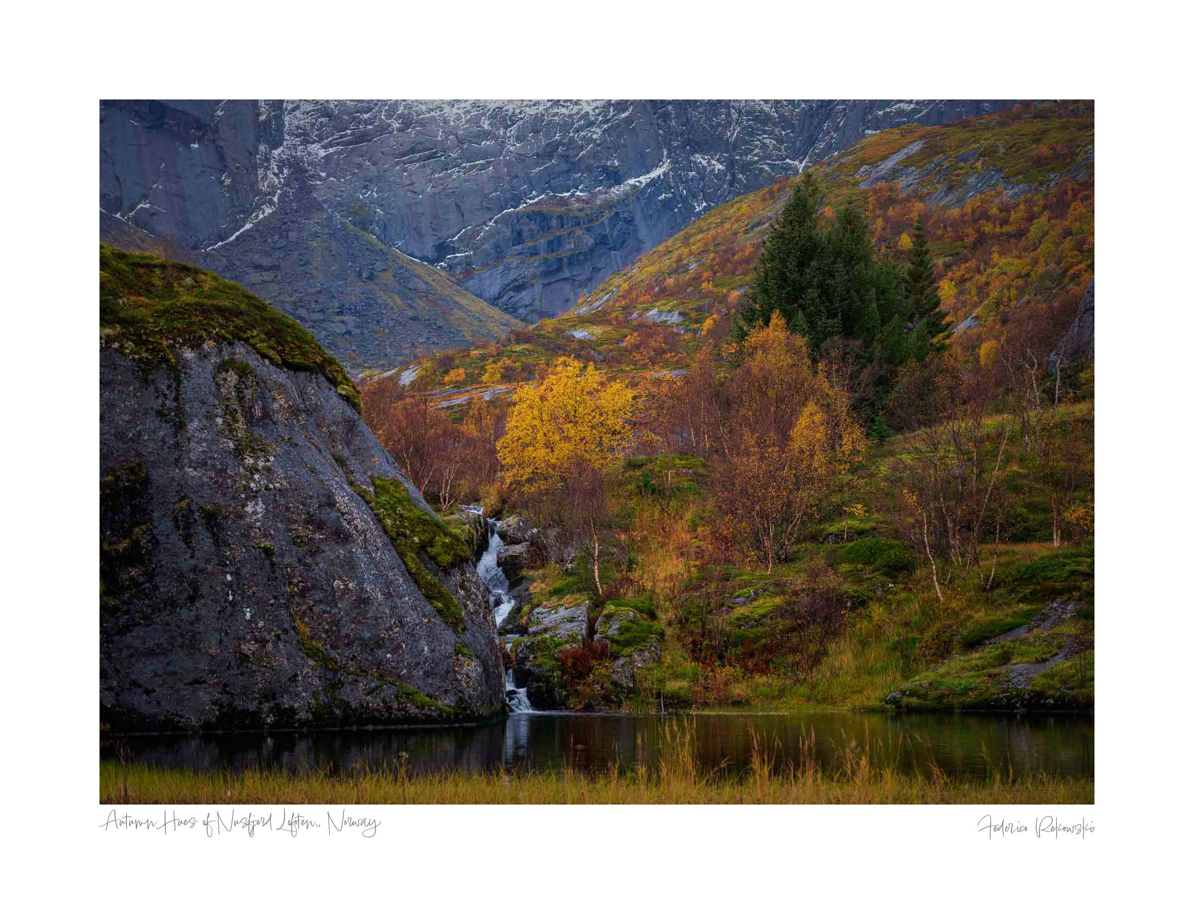 Small waterfall flowing into a pond amidst colorful autumn trees in Nusfjord, Lofoten, with rugged cliffs in the background.