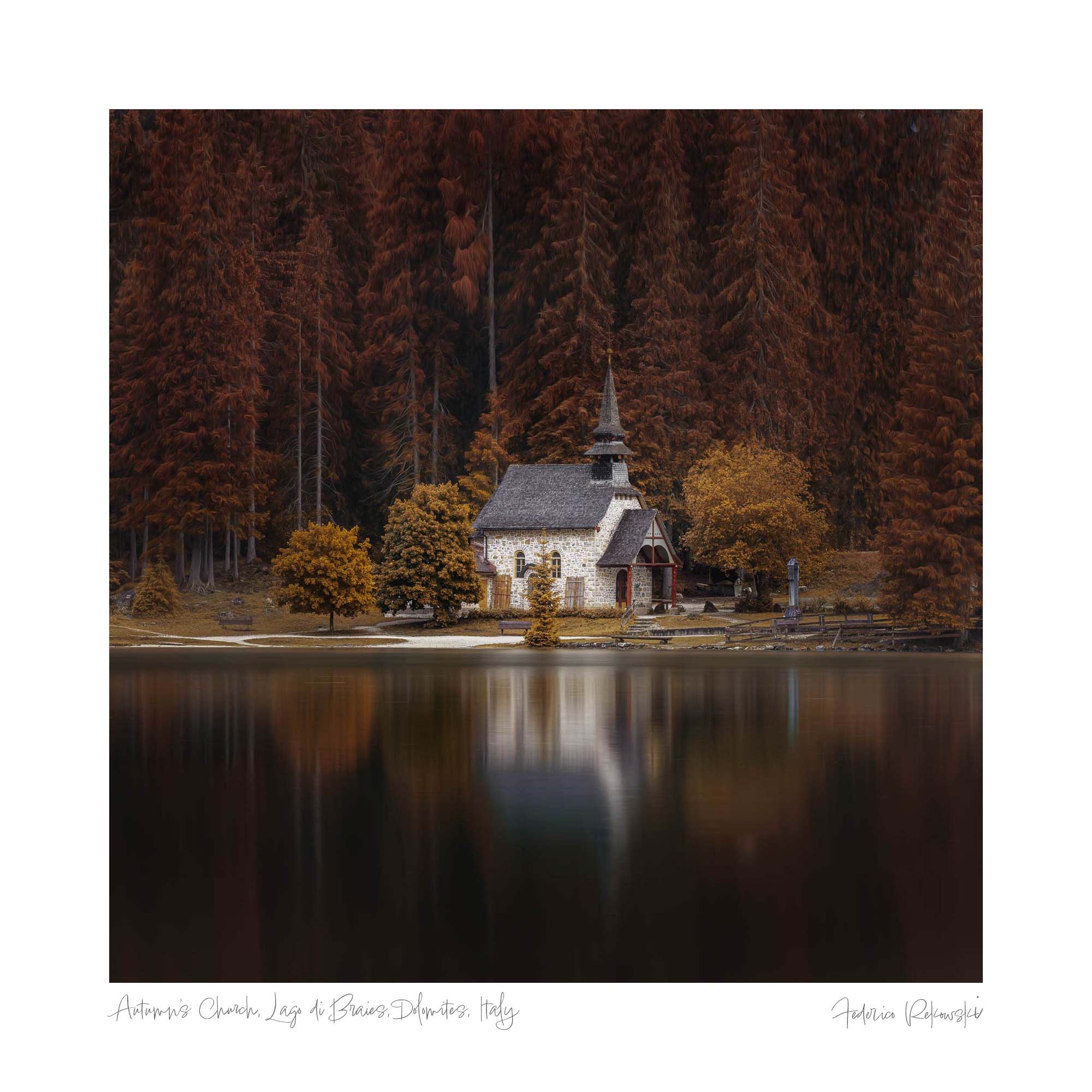 A small stone chapel with a spire, surrounded by trees in autumn colors and reflected in the calm waters of Lago di Braies, Dolomites.