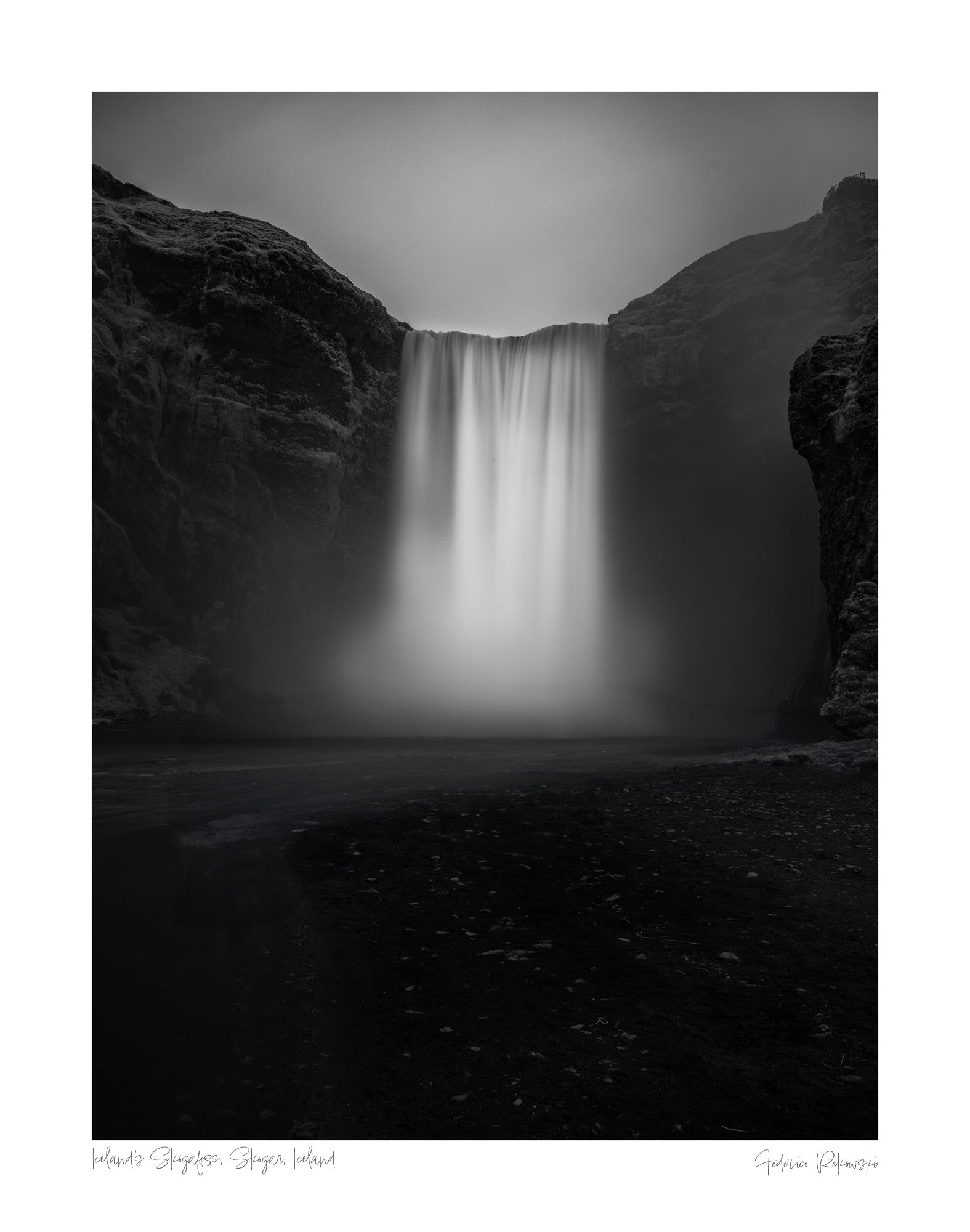 A long exposure photograph of Skógafoss waterfall in Iceland, rendered in stunning black and white, capturing the smooth flow of water between two dark cliffs.