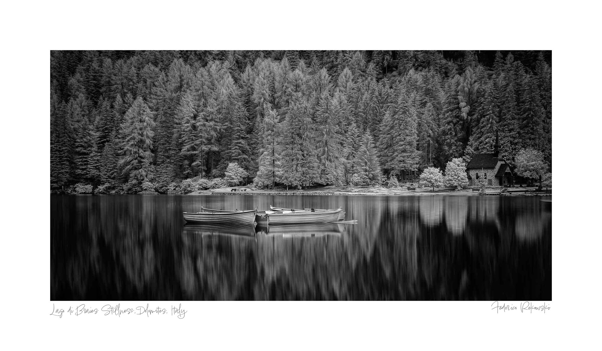 Black and white image of Lago di Braies with still water, reflective boats, a small chapel, and dense trees in the background.