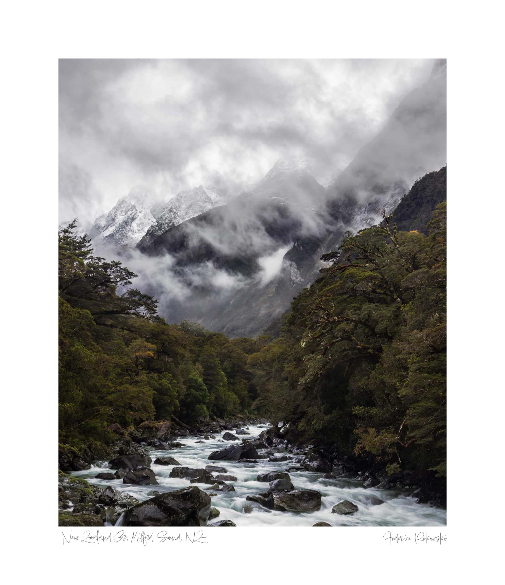 A powerful river cuts through a verdant forest with snow-covered mountains enshrouded in mist in the background at Milford Sound, New Zealand.