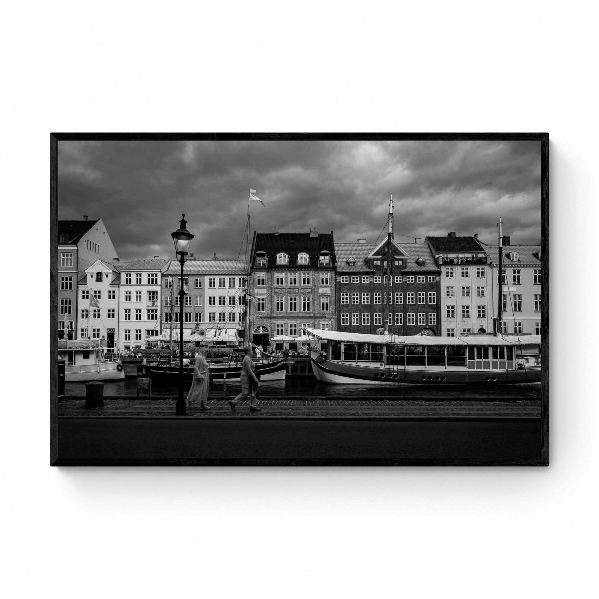 Black and white image of Nyhavn, Copenhagen, with historical buildings along the waterfront, boats docked in the canal, and people walking by.