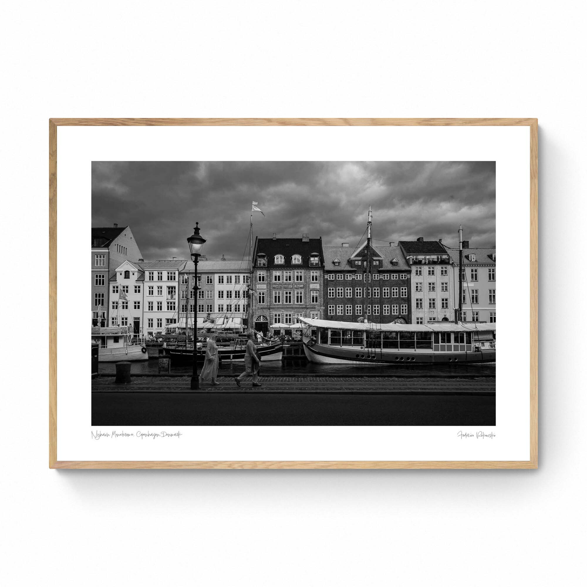 Black and white image of Nyhavn, Copenhagen, with historical buildings along the waterfront, boats docked in the canal, and people walking by.