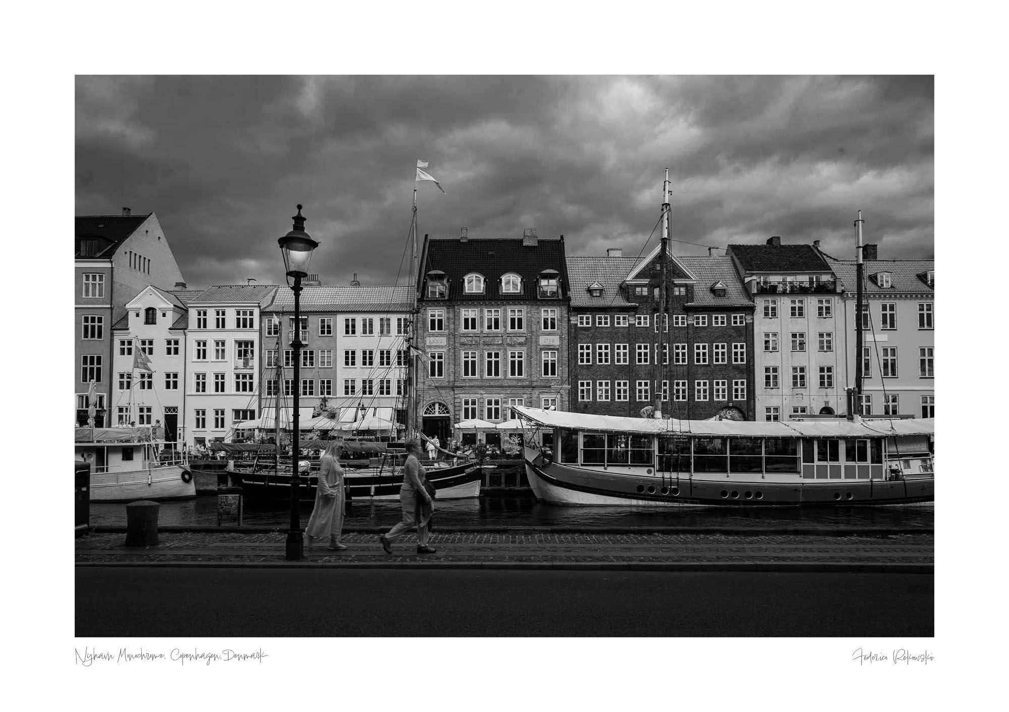 Black and white image of Nyhavn, Copenhagen, with historical buildings along the waterfront, boats docked in the canal, and people walking by.