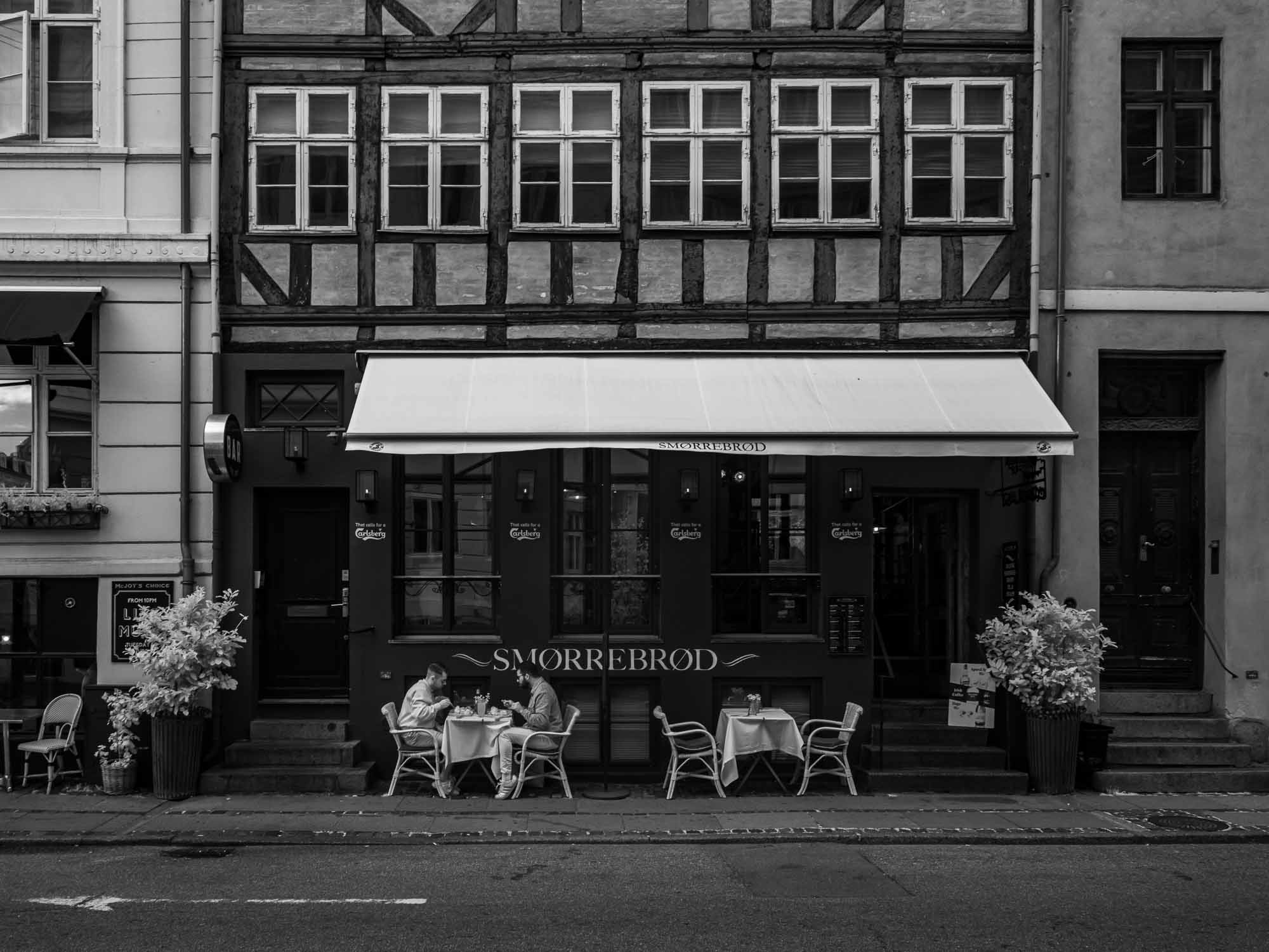 Black and white photo of a traditional Danish Smørrebrød café in Nyhavn, Copenhagen, with outdoor seating and historical buildings.