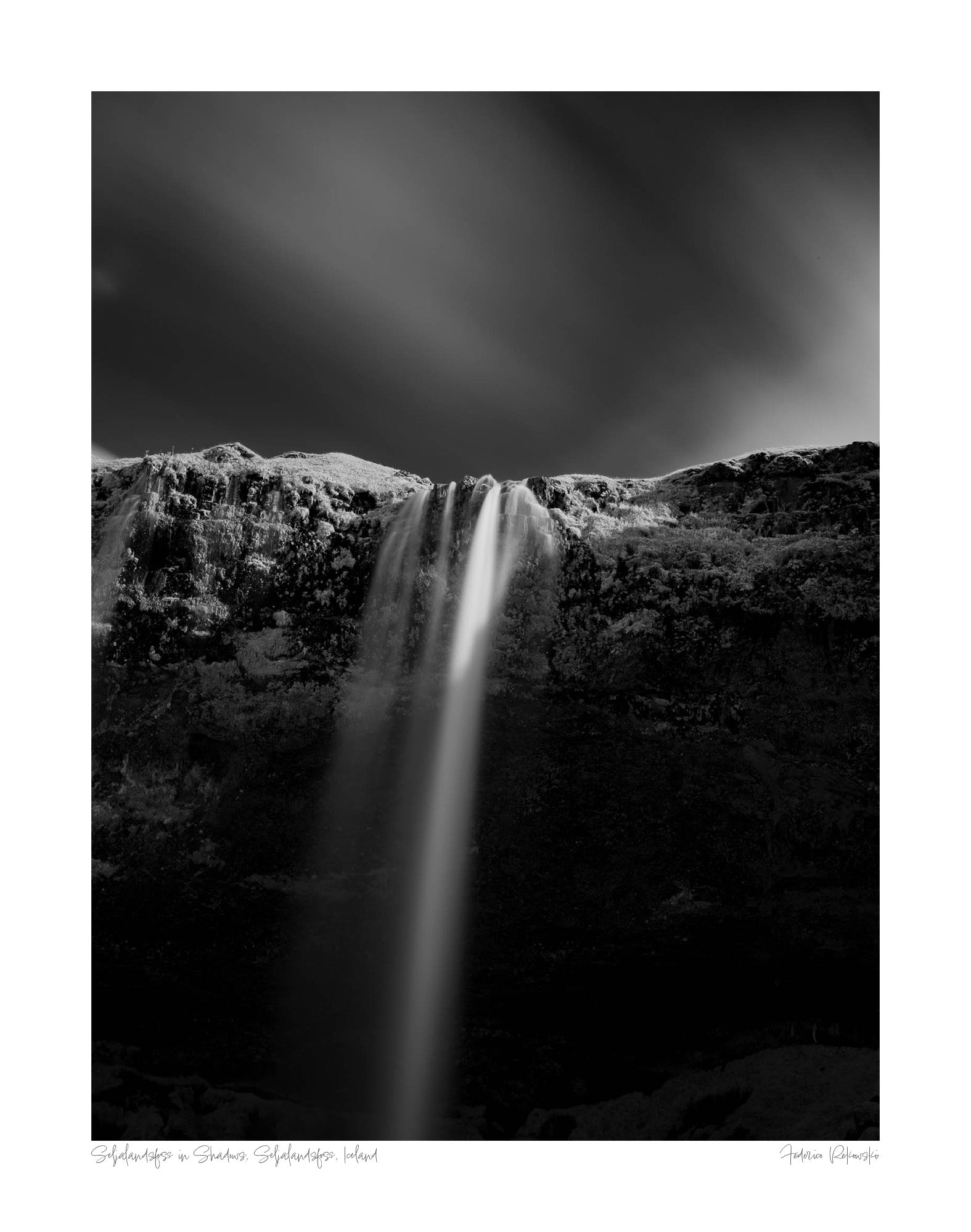 A black and white long-exposure photograph of Seljalandsfoss waterfall in Iceland, showing smooth water streams against a dark, textured cliff.