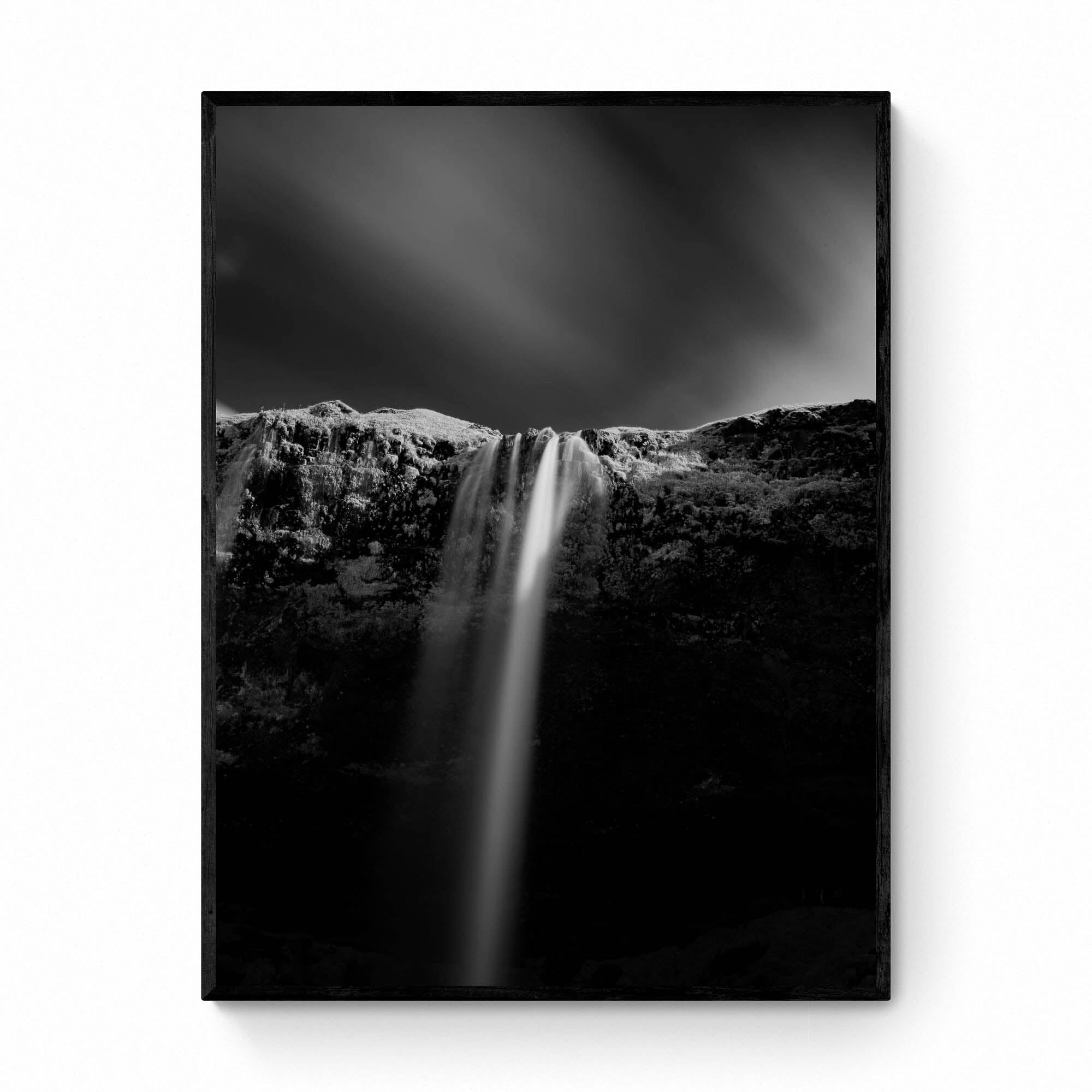 A black and white long-exposure photograph of Seljalandsfoss waterfall in Iceland, showing smooth water streams against a dark, textured cliff.