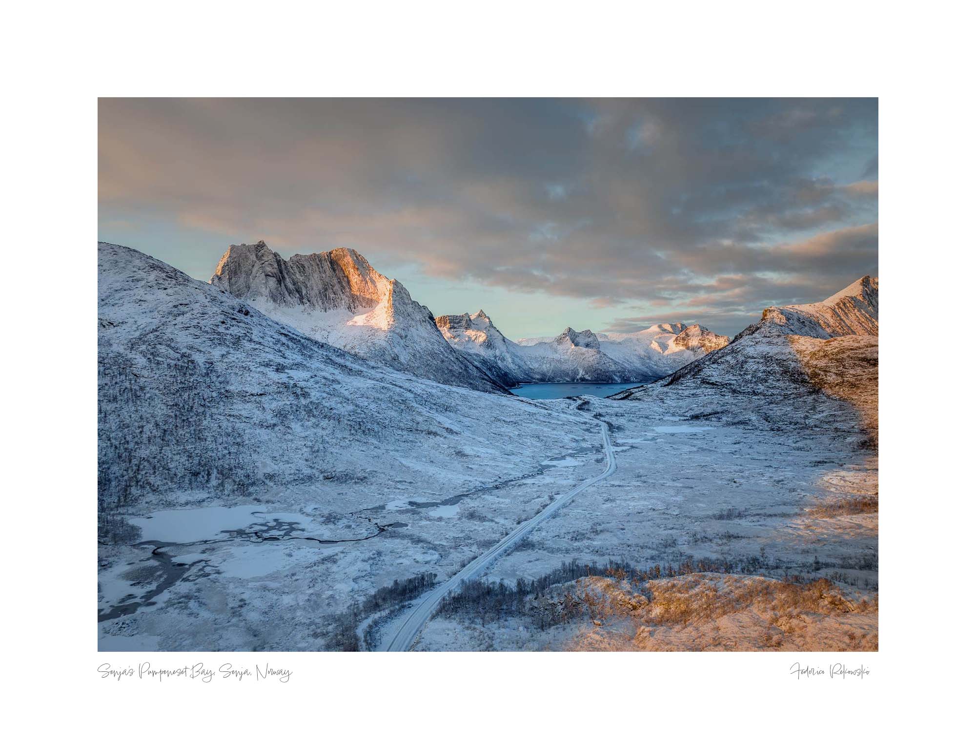 Snow-covered landscape at Husøy, Senja, with mountains glowing in the soft light of the setting sun and a road winding through the valley.