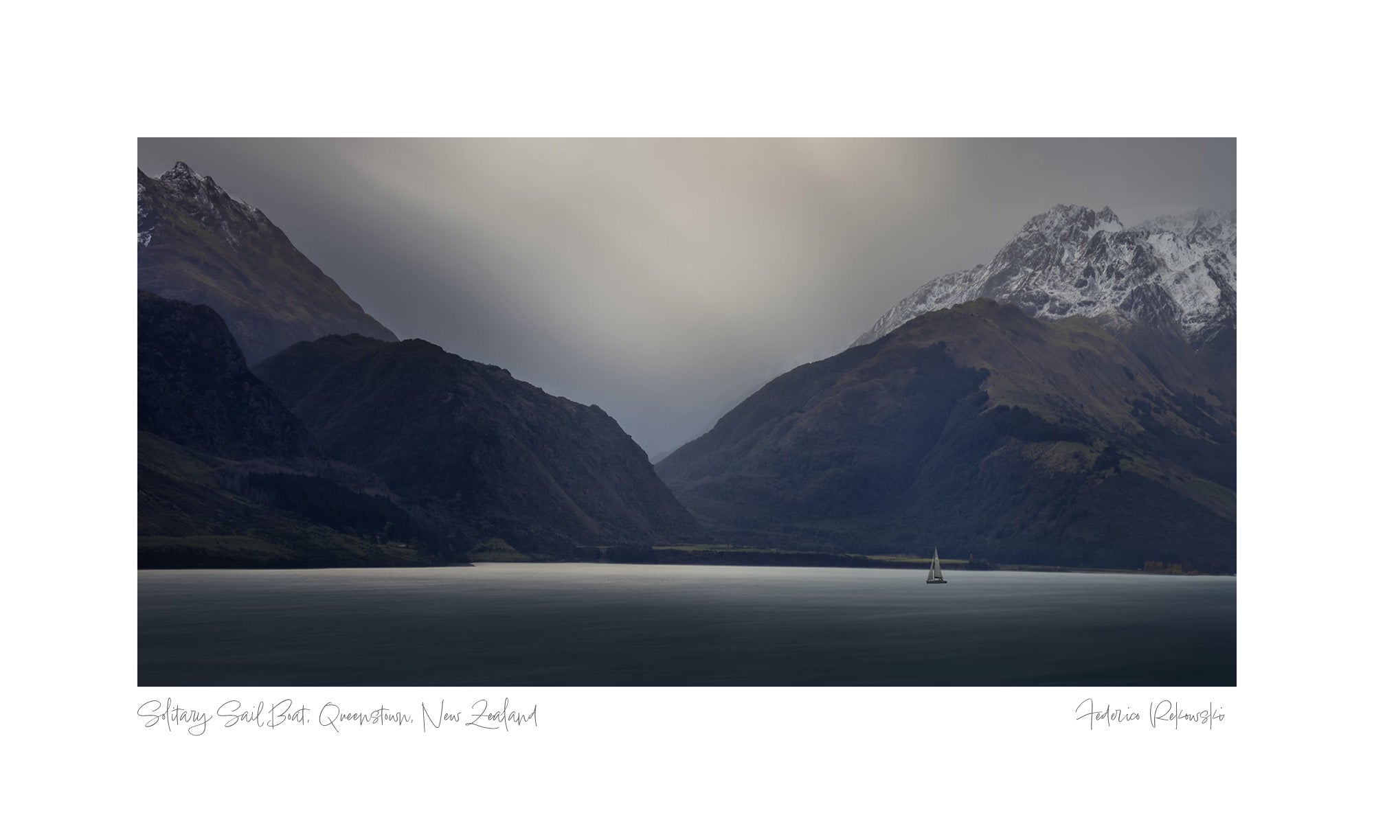 A lone sailboat on a calm lake in Queenstown, New Zealand, with towering, snow-capped mountains under a soft, overcast sky.