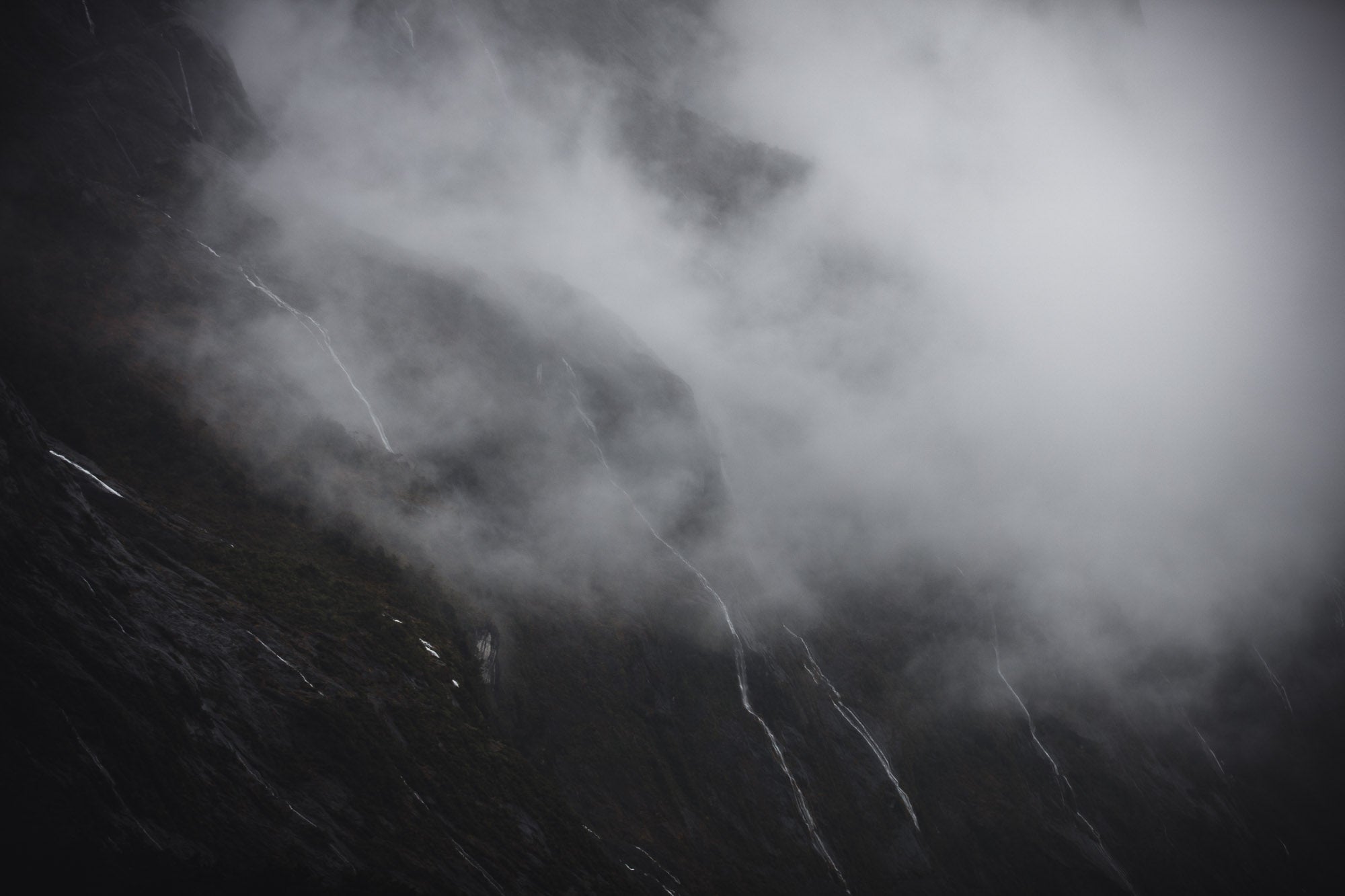 Photograph titled 'Tears of the Sound' showing misty waterfalls on the cliffs of Milford Sound, New Zealand, resembling tears against a somber backdrop.