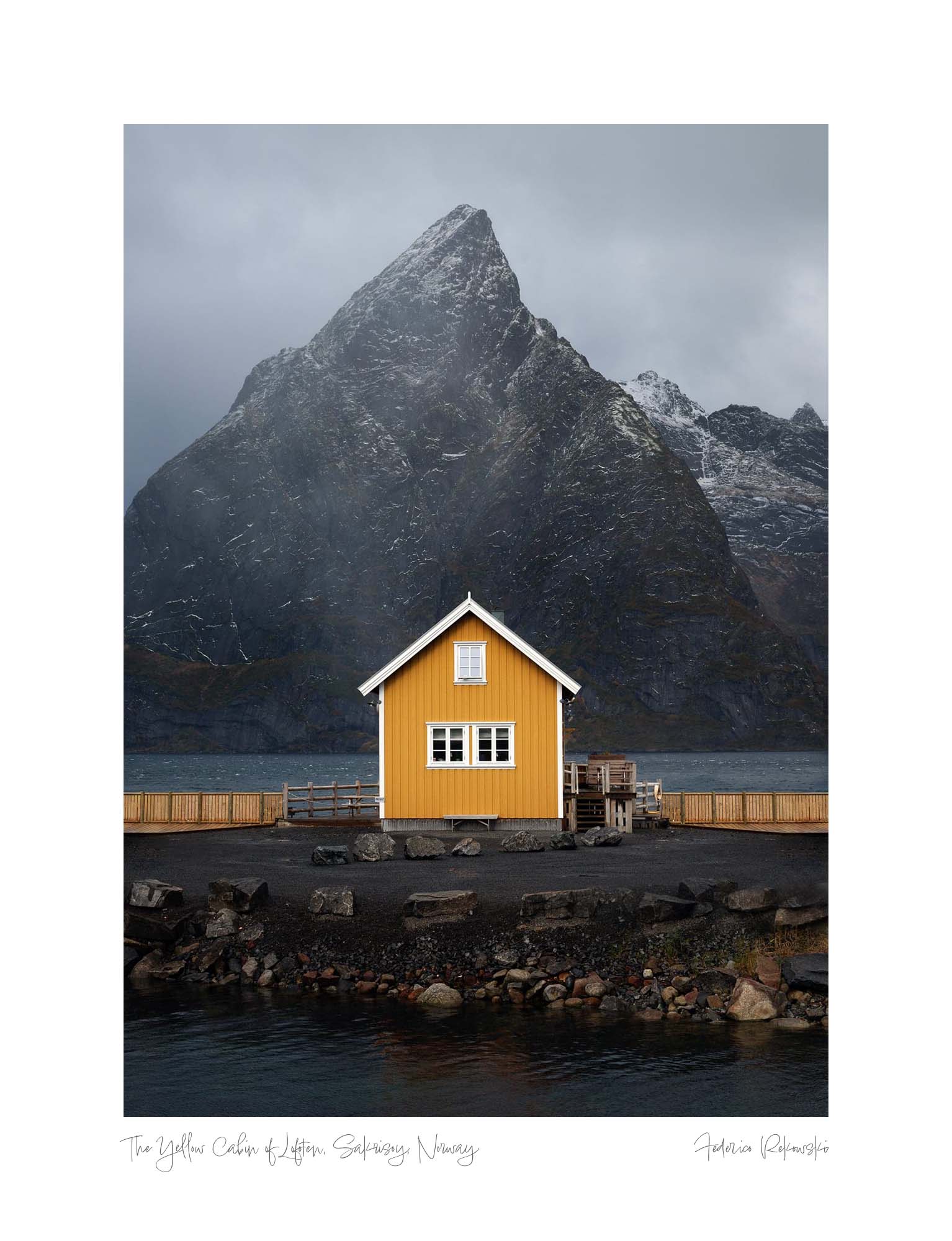 A solitary yellow cabin stands in the foreground with a towering, snow-streaked mountain rising dramatically behind it in Lofoten, Norway. Wall Art