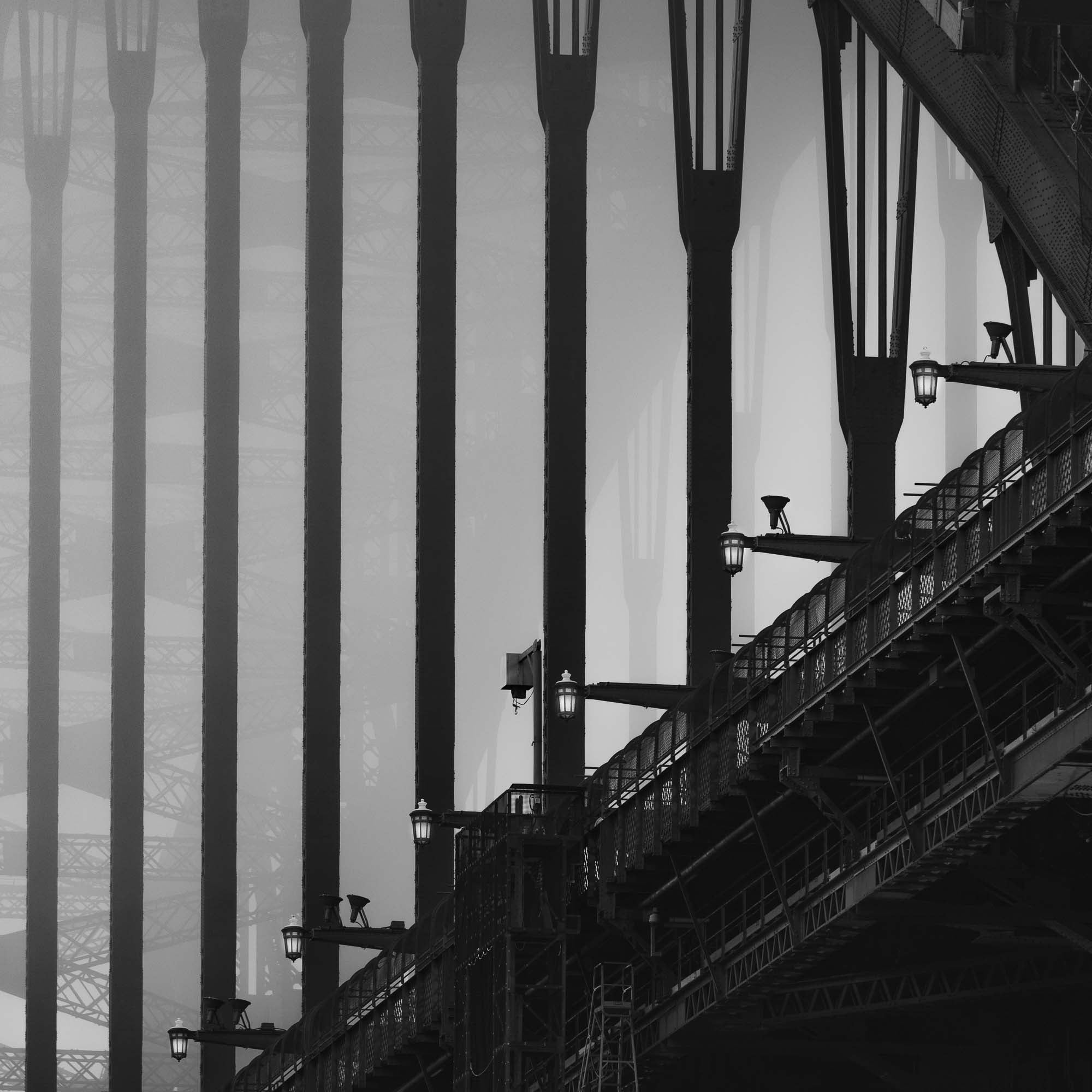 Monochrome image of Sydney Harbour Bridge, showing the contrast of the stark, strong lines of its arches and street lamps against the light sky.